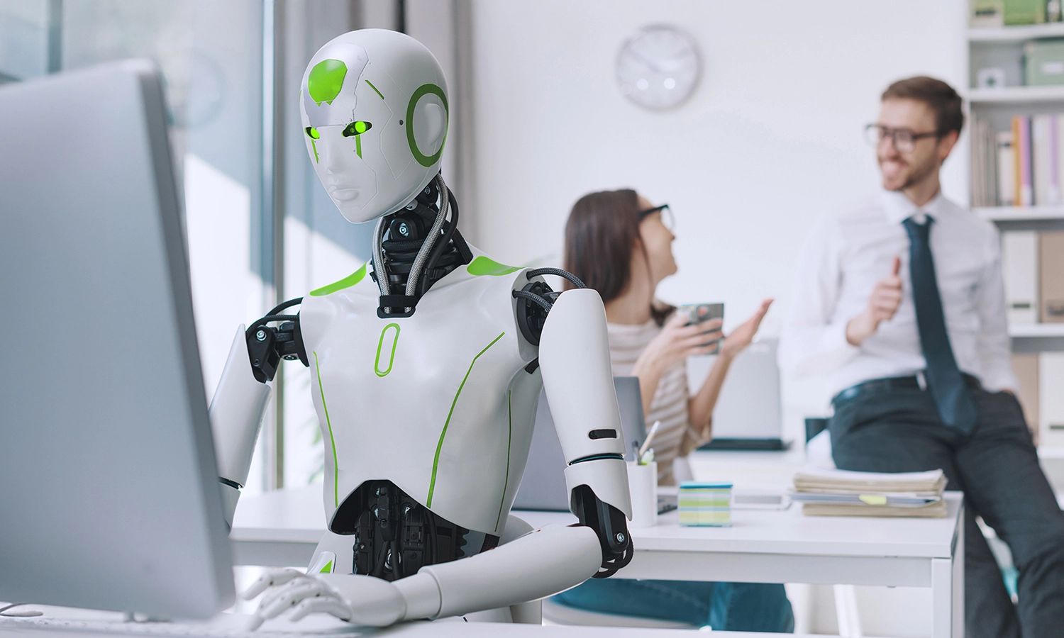 Humanoider Roboter arbeitet am Computer in einem modernen Büro, während zwei Kollegen im Hintergrund miteinander sprechen – Symbolbild für Zusammenarbeit zwischen Mensch und künstlicher Intelligenz.