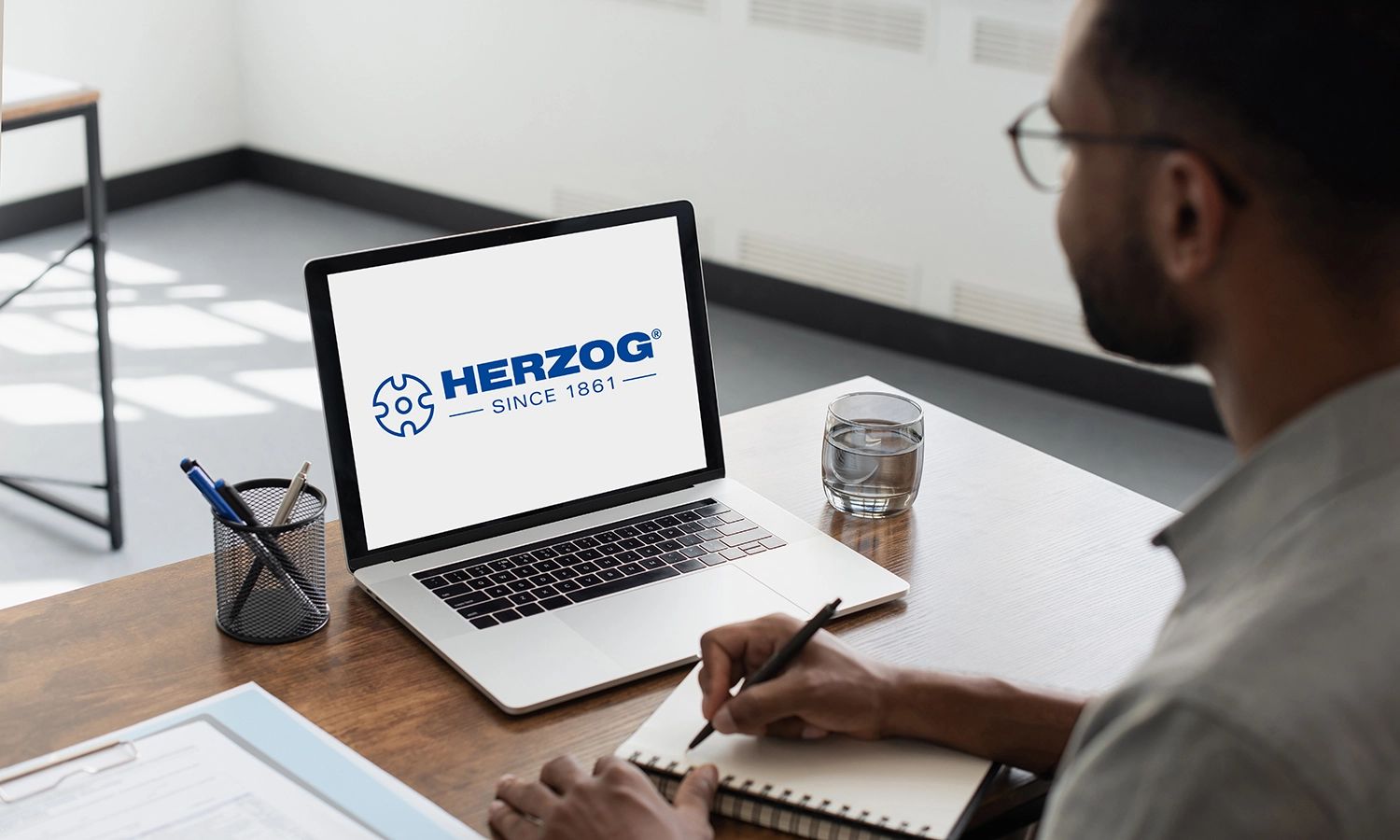 Person sitzt an einem Schreibtisch und schaut auf einen Laptop, auf dessen Bildschirm das blaue Logo von HERZOG mit dem Schriftzug ‚HERZOG – Since 1861‘ und einem stilisierten Zahnrad-Symbol angezeigt wird.