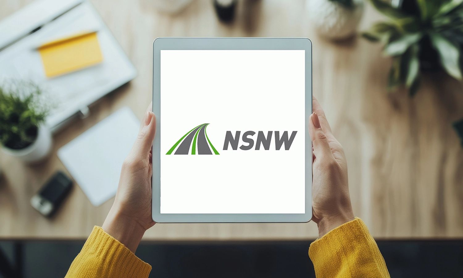 Person hält ein Tablet, auf dessen Bildschirm das Logo von NSNW mit grauem Schriftzug und einer stilisierten grünen und grauen Straßen-Grafik angezeigt wird.
