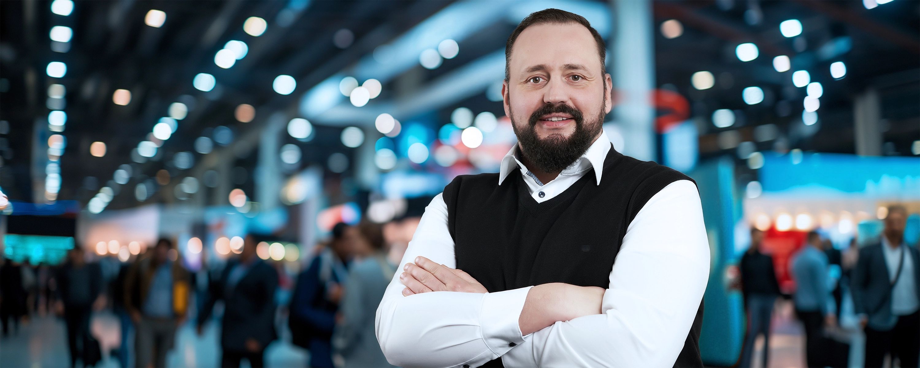 Ein Mann mittleren Alters mit dunklem Bart und einem weißen Hemd mit schwarzer Weste steht mit verschränkten Armen und einem Lächeln in einer unscharfen, belebten Messehalle oder Veranstaltungsumgebung.