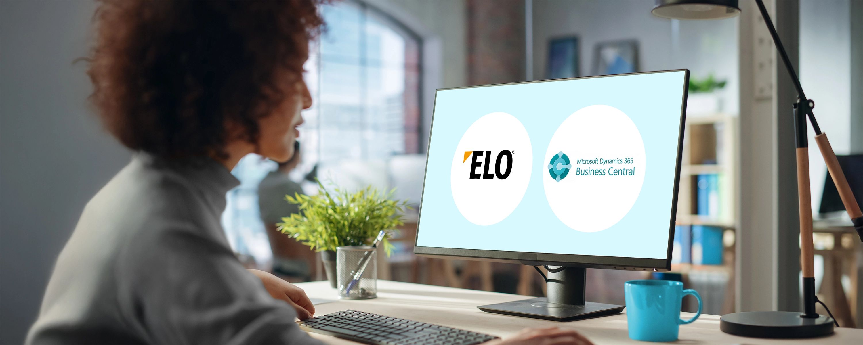 Eine Frau mit lockigen Haaren sitzt in einem modernen Büro an einem Schreibtisch und tippt. Auf dem Computerbildschirm vor ihr sind die Logos von "ELO" und "Microsoft Dynamics 365 Business Central nebeneinander dargestellt.