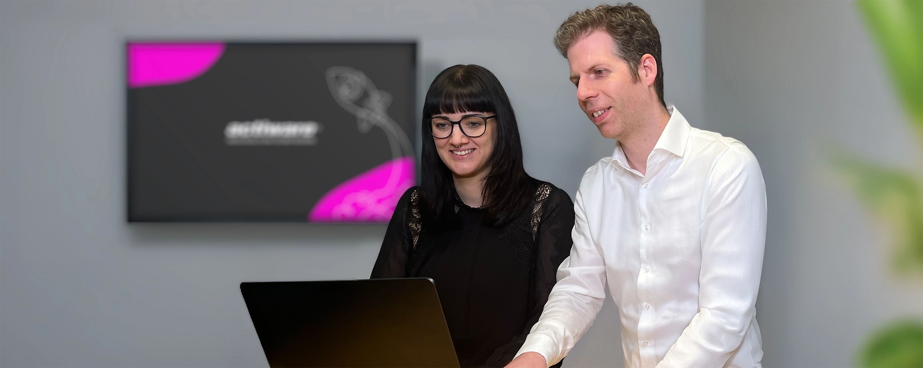Zwei Mitarbeitende arbeiten zusammen, im Hintergrund hängt ein Bildschirm mit ACTIWARE-Branding.