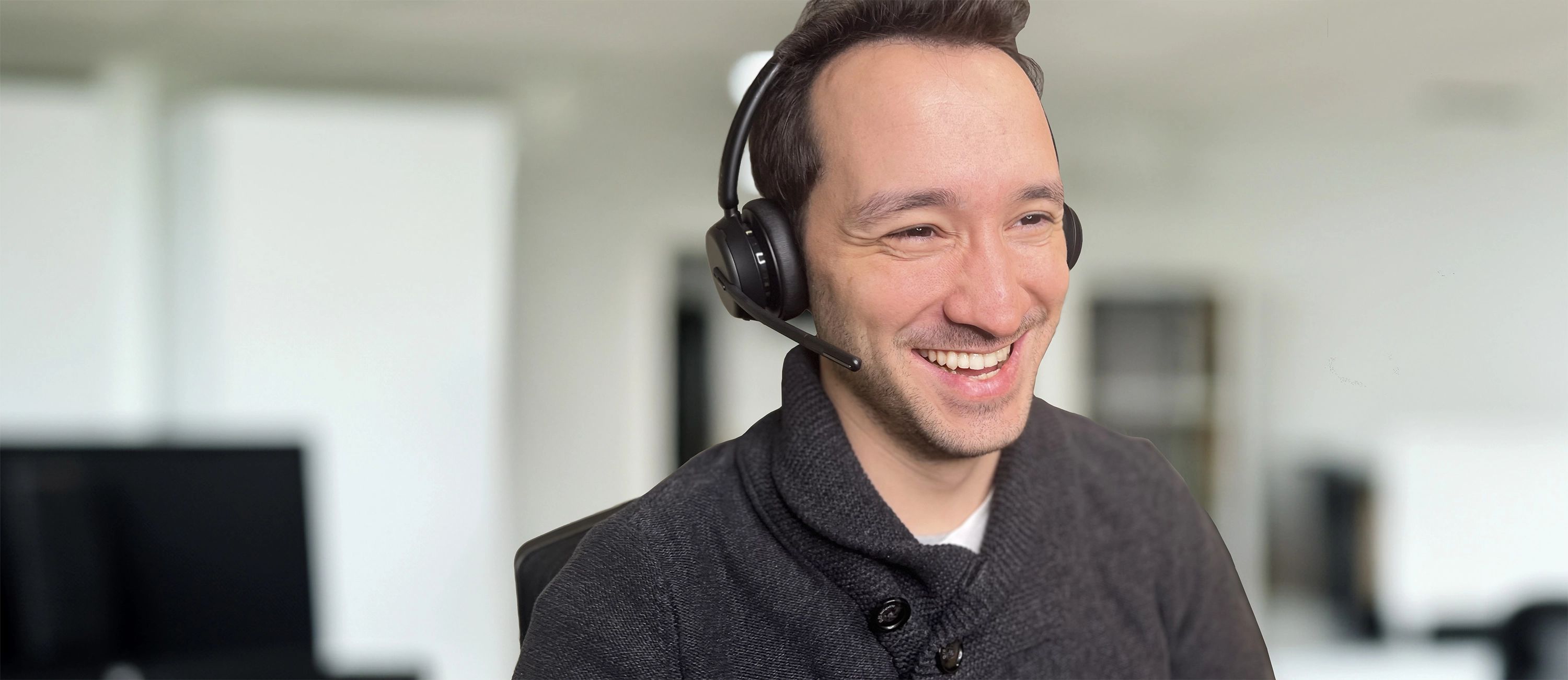 Mann mit Headset arbeitet online und lächelt vor einem Bürohintergrund.