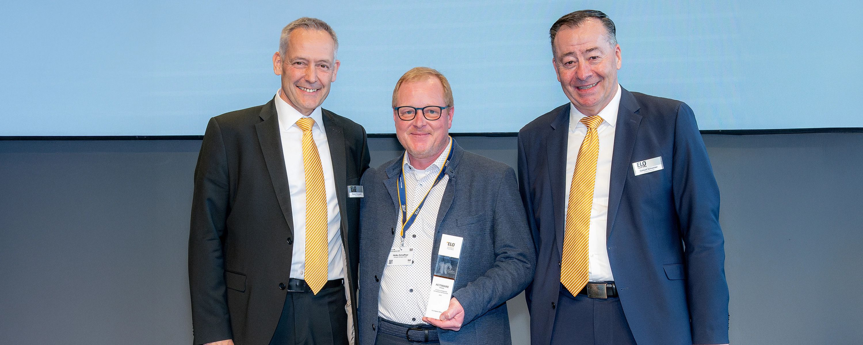 Drei Männer in Anzügen posieren für ein Foto bei einer Preisverleihung. Der Mann in der Mitte ist Heiko Schaffner, CEO der ACTIWARE Schweiz GmbH, und hält stolz eine Trophäe, während die beiden Männer an seiner Seite, erkennbar an ihren gelben Krawatten und ELO-Namensschildern, freundlich lächeln.