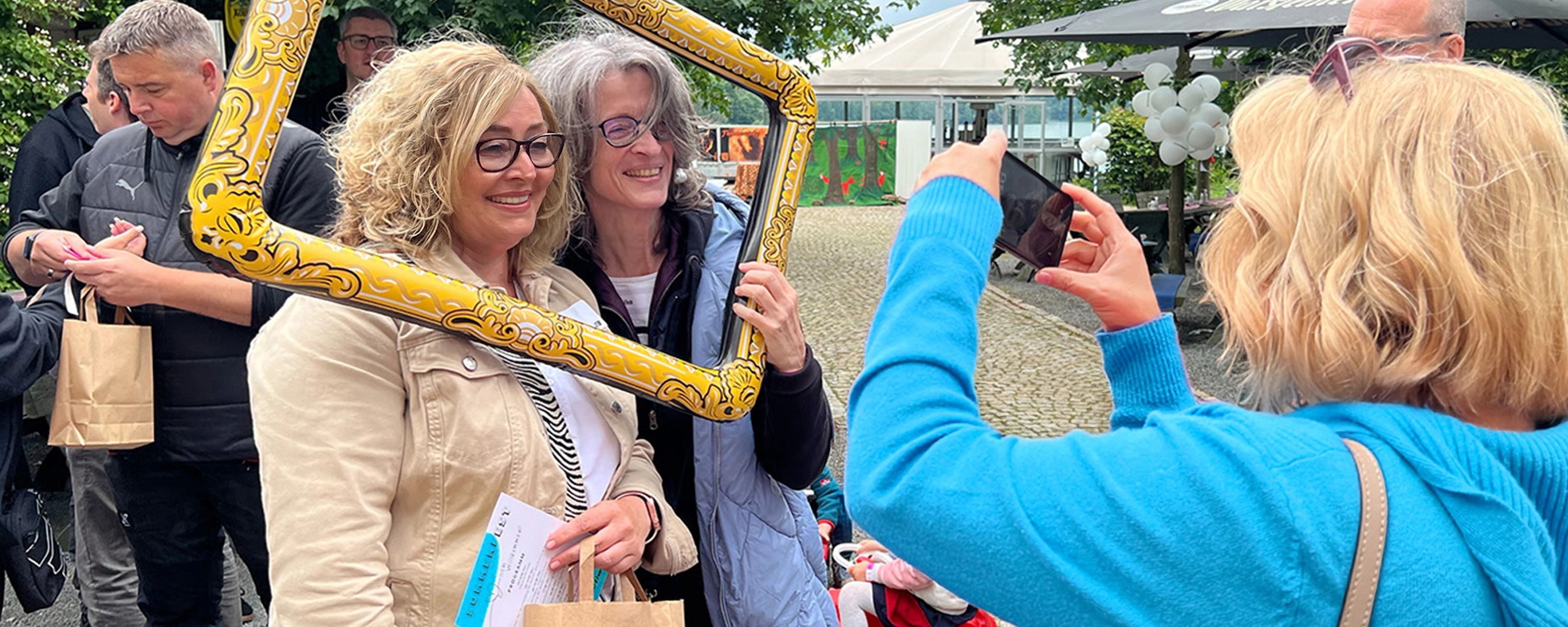 Eine Frau in einem blauen Pullover fotografiert mit ihrem Smartphone zwei lächelnde Frauen, die bei einer Veranstaltung im Freien gemeinsam in einem großen, gelben Bilderrahmen posieren.