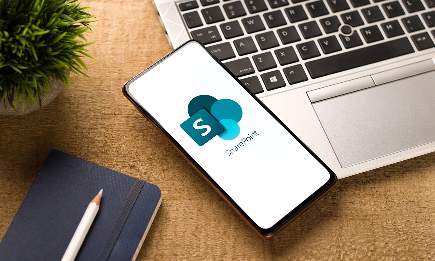 Smartphone mit dem Microsoft SharePoint Logo auf dem Display, liegend auf einem Laptop neben einem Notizbuch und einer Pflanze.