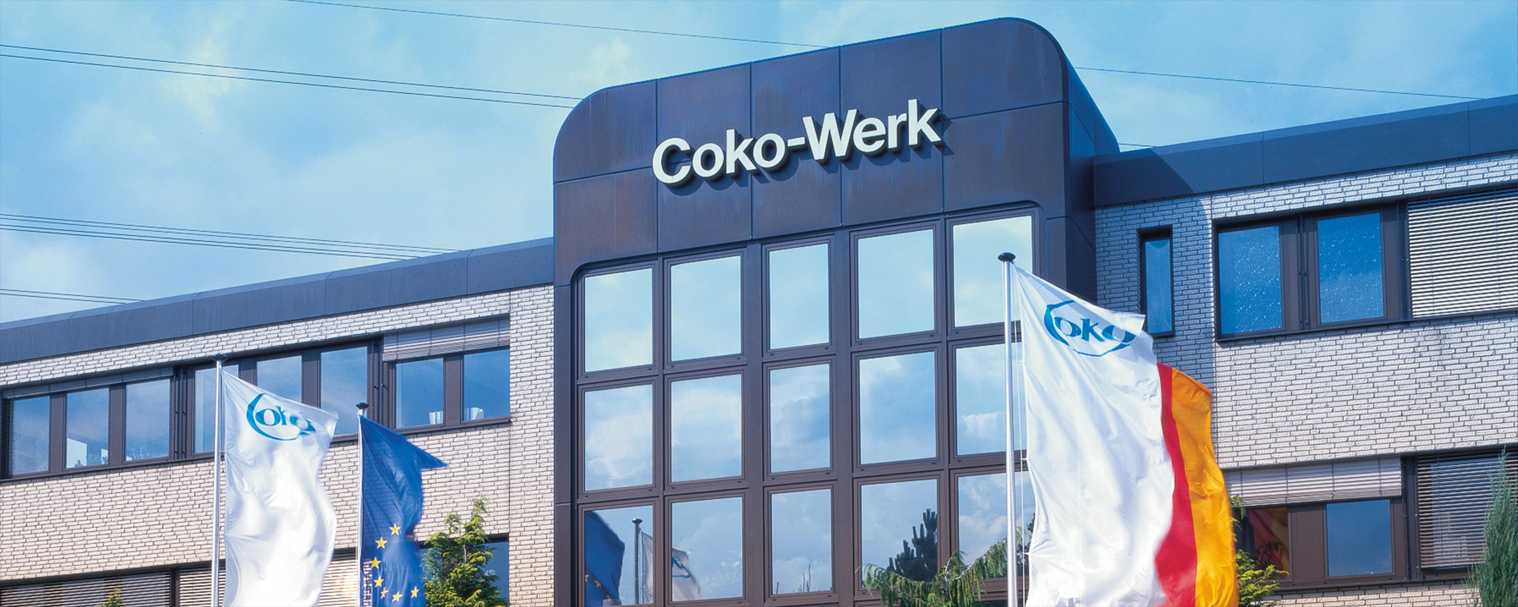 Moderne Firmenzentrale des Coko-Werks mit Unternehmens- und Landesflaggen – führender Hersteller in der Kunststoffverarbeitung und industriellen Produktion.