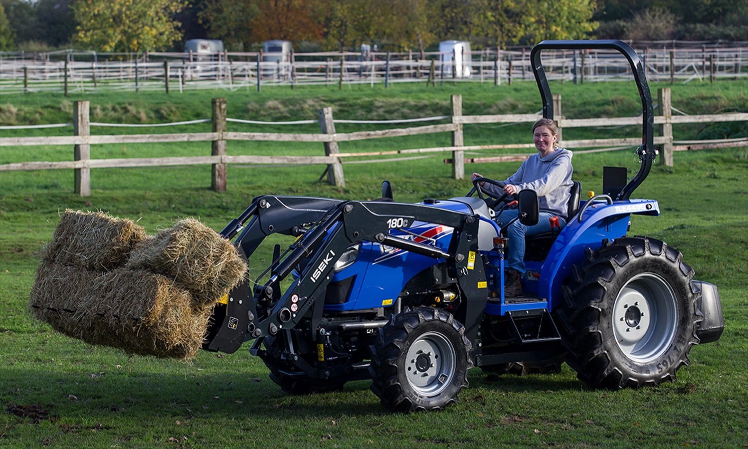 Iseki‑Traktor mit Frontlader transportiert Heuballen auf einer Weide – vielseitige Landmaschine für Landwirtschaft, Tierhaltung und Geländearbeiten.