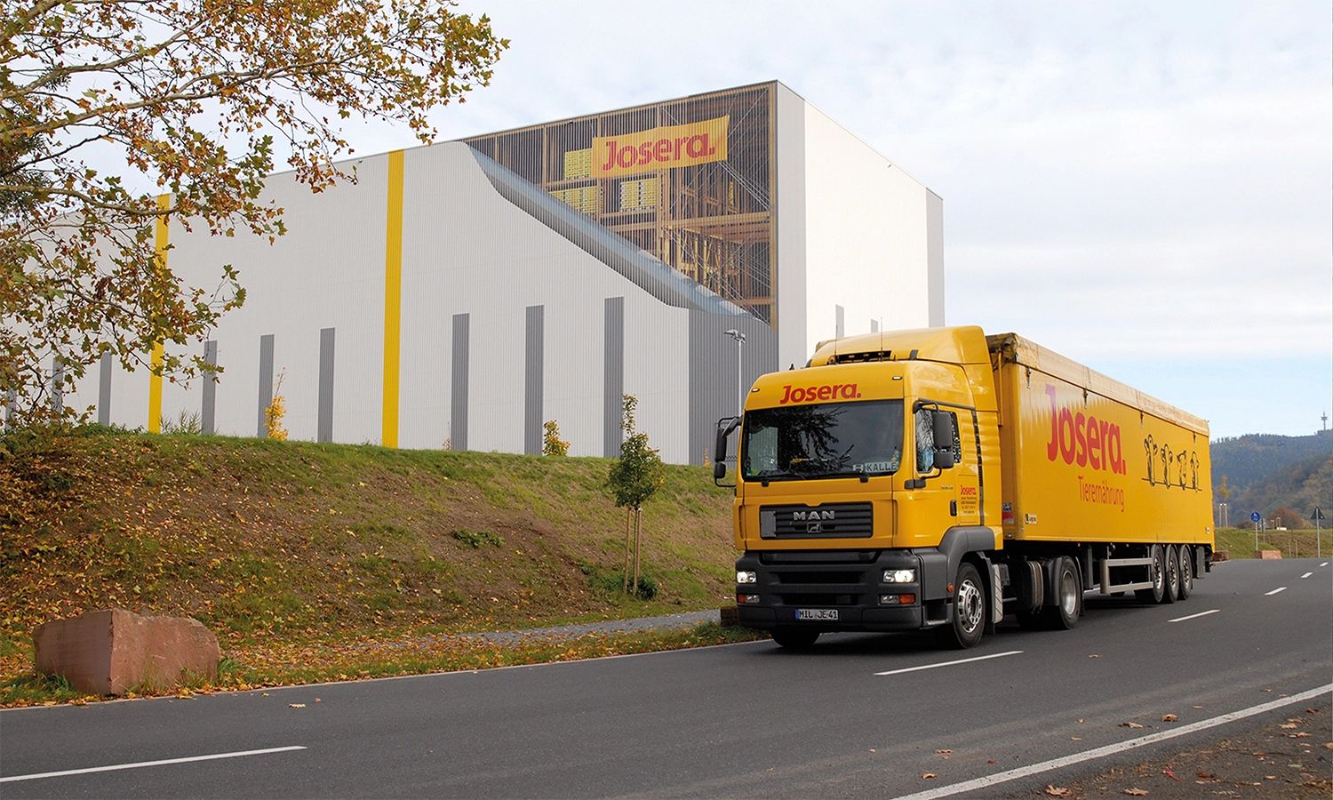 Josera‑Lkw fährt am Firmengebäude vorbei – moderner Produktionsstandort und Logistiktransport für Tiernahrung und landwirtschaftliche Produkte.
