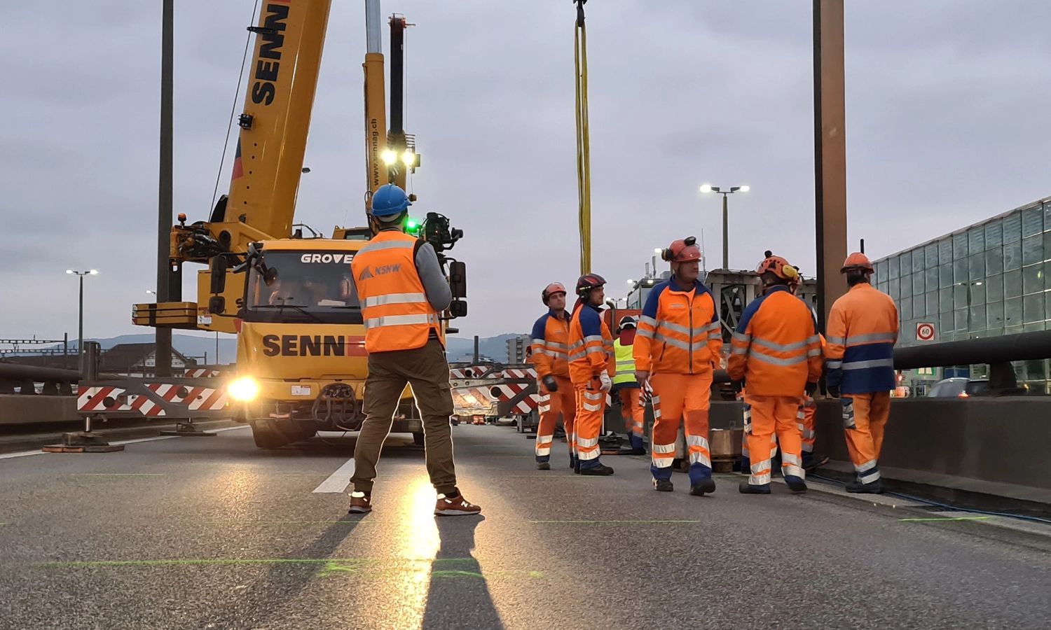 NSNW‑Bauteam bei Brückenarbeiten – Straßen‑ und Tiefbauarbeiter in Sicherheitskleidung koordinieren Hebeeinsatz mit Kran auf Baustelle.