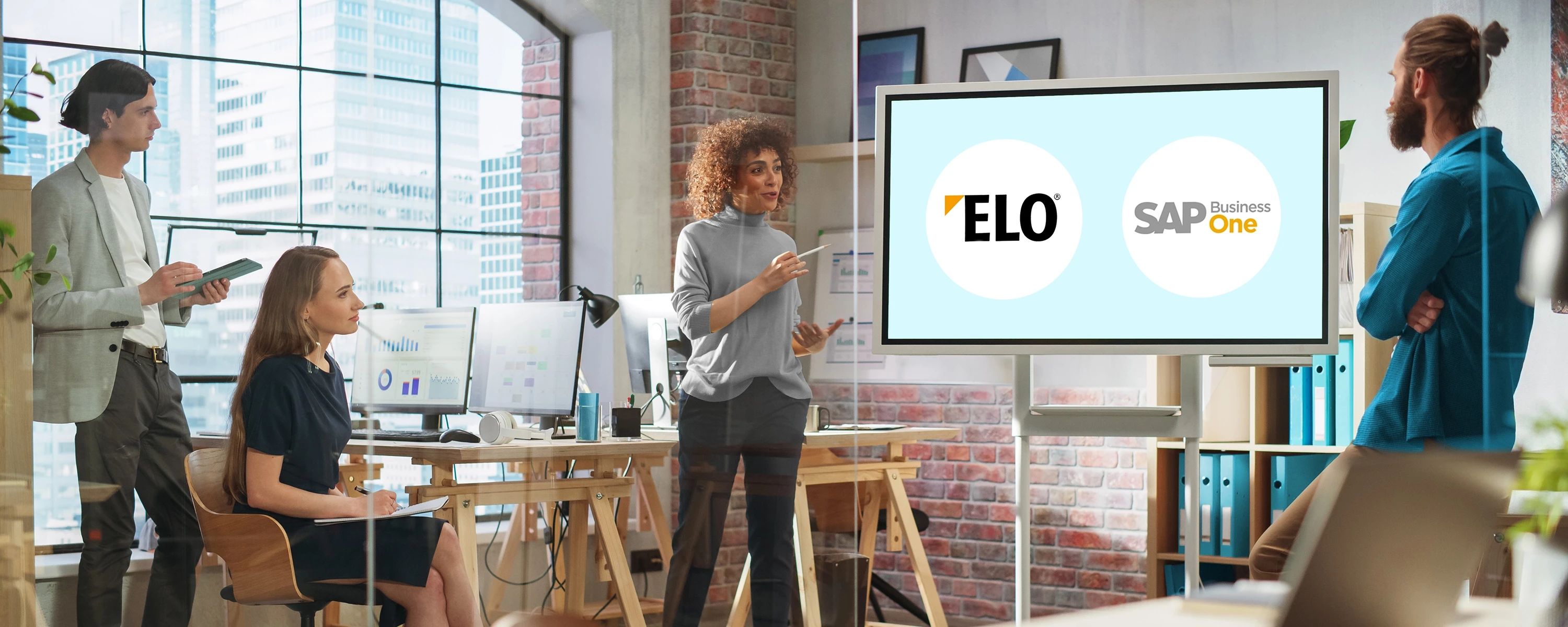 Eine Gruppe von vier Personen in einem modernen Büro hält eine Präsentation ab. Eine Frau steht in der Mitte und präsentiert auf einem großen Bildschirm die Logos von "ELO" und "SAP Business One".