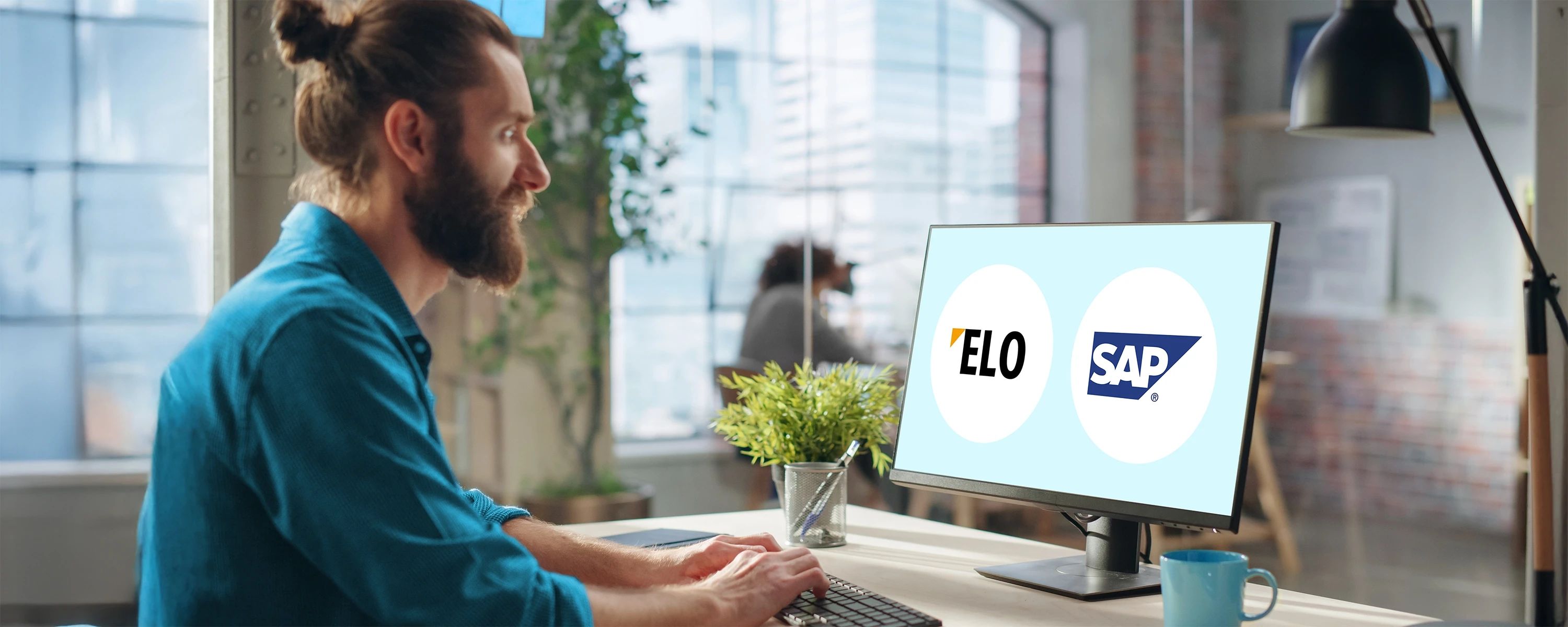 Ein Mann mit einem blauen Hemd, einem Bart und einem Dutt sitzt in einem modernen Büro vor einem Computerbildschirm. Der Bildschirm zeigt die Logos von "ELO" und "SAP" nebeneinander.