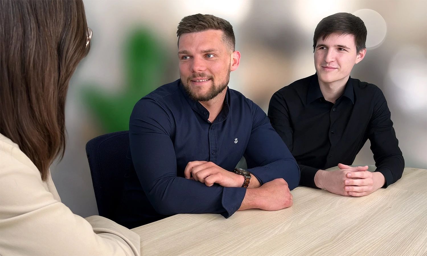 Drei Personen sitzen an einem hellen Holztisch und sind in ein Gespräch vertieft, das Wissenstransfer oder Beratung symbolisiert. Zwei Männer sitzen lächelnd dem Gesprächspartner (einer Frau, deren Rücken zu sehen ist) gegenüber.