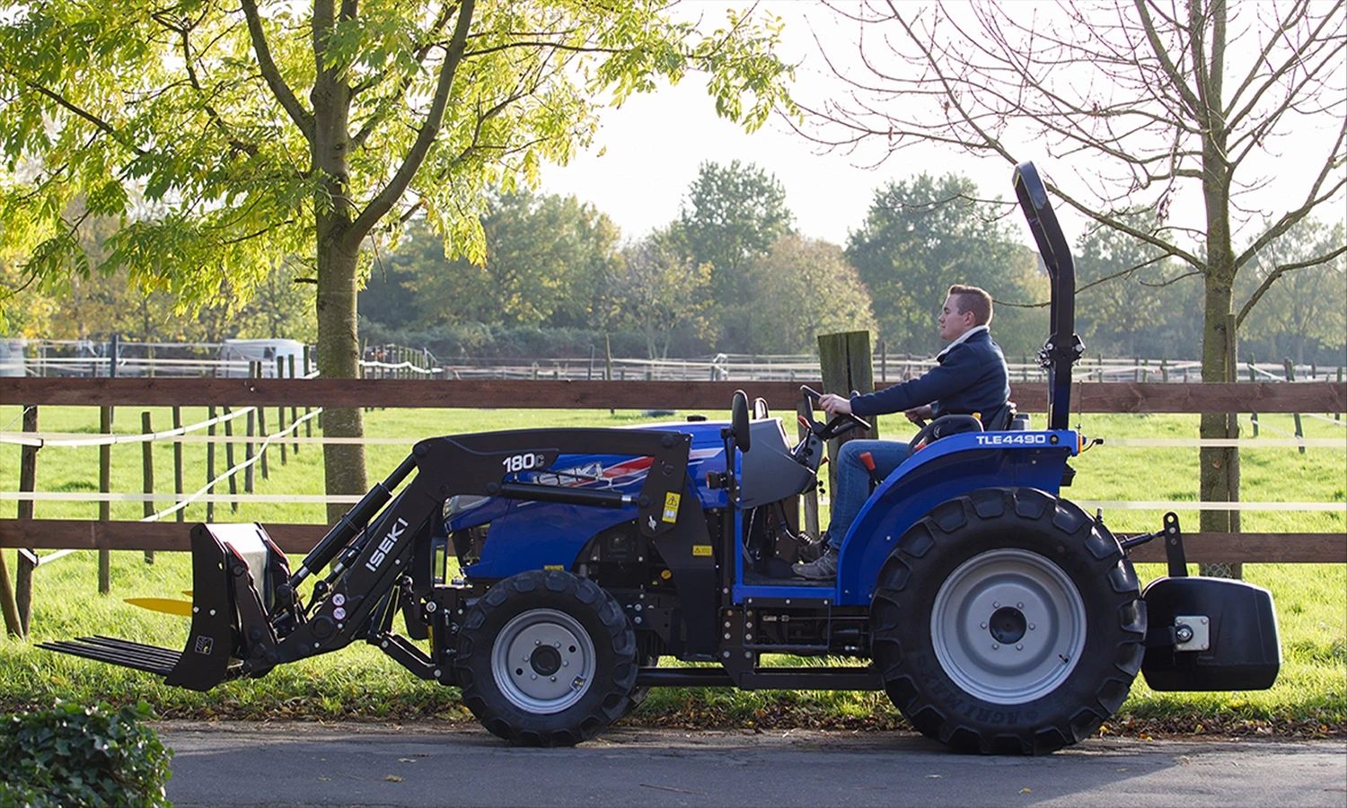 Iseki‑Traktor im Einsatz neben einer Grünfläche – Fahrer bedient blauen Kommunaltraktor TLE4450 mit Frontlader, entwickelt für Landwirtschaft- und Geländepflege.