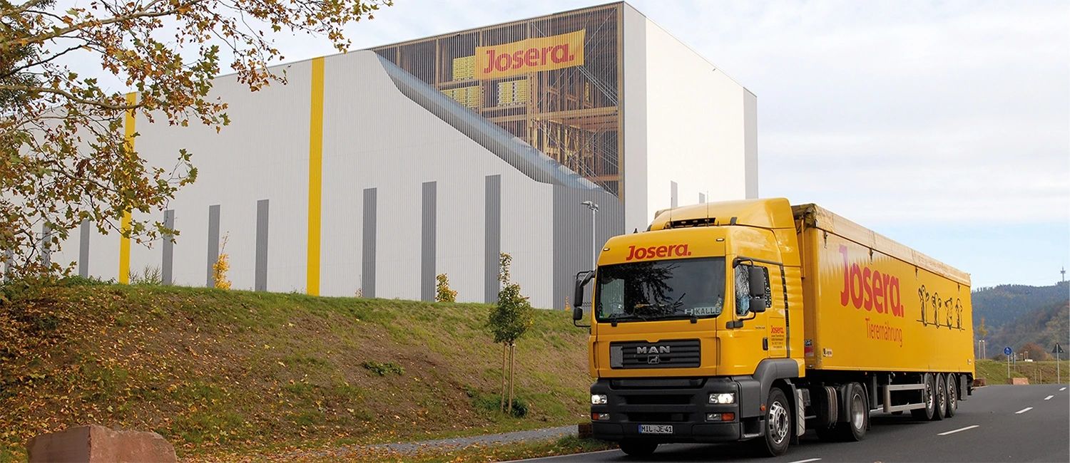 Josera‑Lkw fährt am Firmengebäude vorbei – moderner Produktionsstandort und Logistiktransport für Tiernahrung und landwirtschaftliche Produkte.