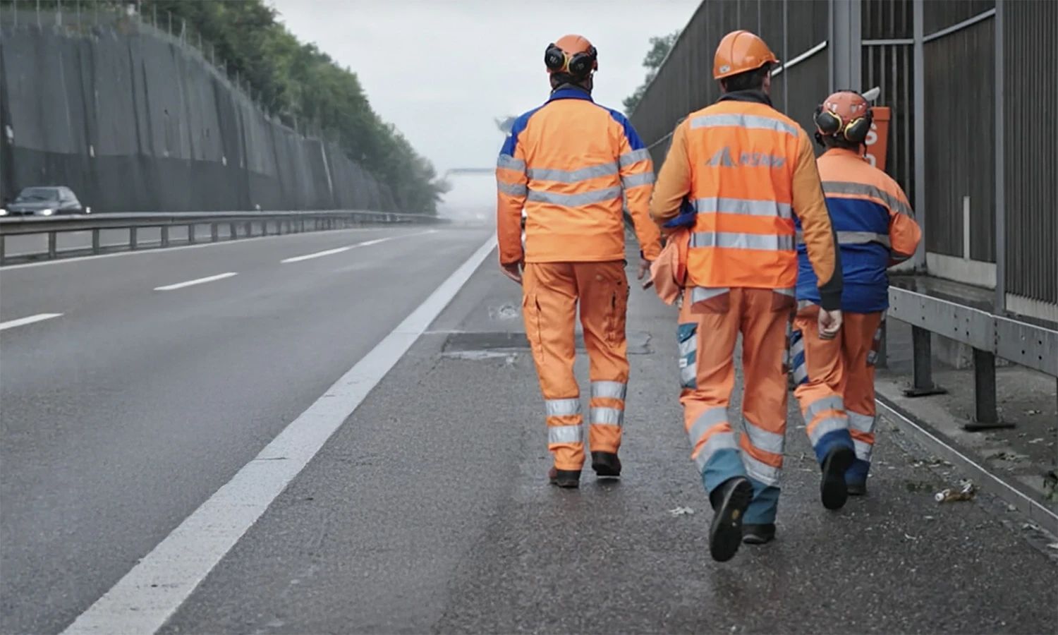 NSNW‑Mitarbeiter in Warnkleidung gehen entlang einer Autobahn – Straßenbau‑ und Instandhaltungsteam bei Arbeiten an Lärmschutzwand.