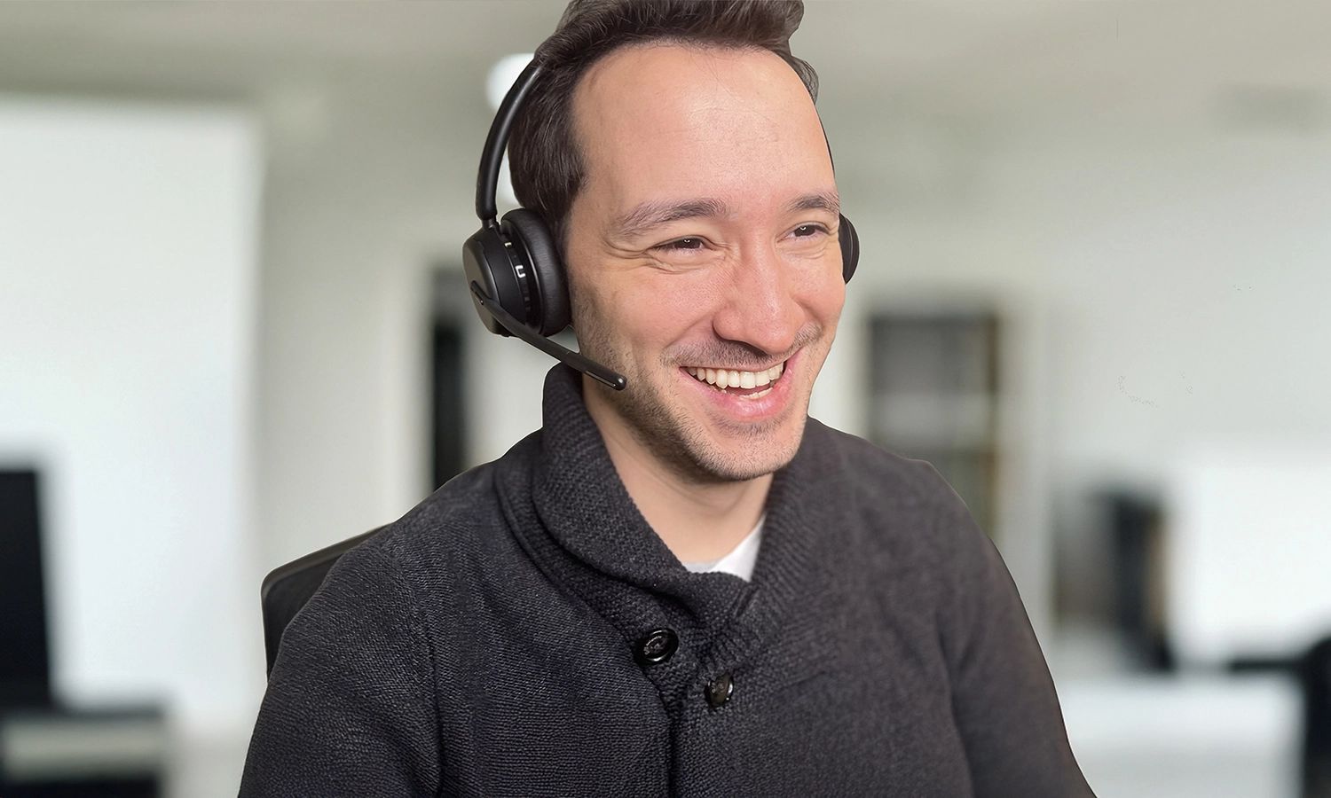 Mann mit Headset arbeitet online und lächelt vor einem Bürohintergrund.