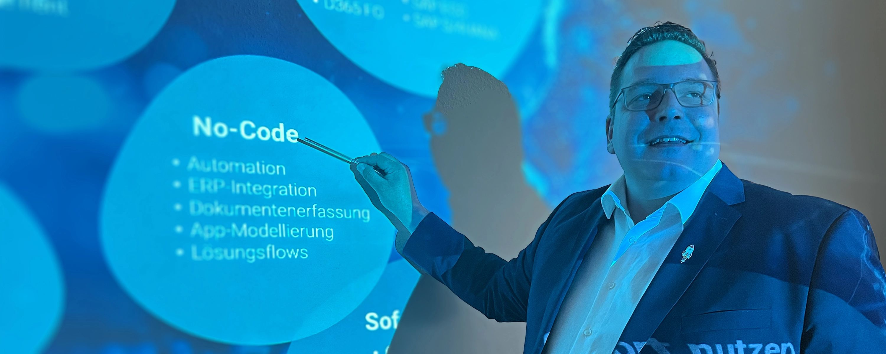 Mann hält eine Schulung ab und zeigt mit einem Stift auf eine blaue Projektion mit dem Titel "No-Code" und einer Liste von Stichpunkten.