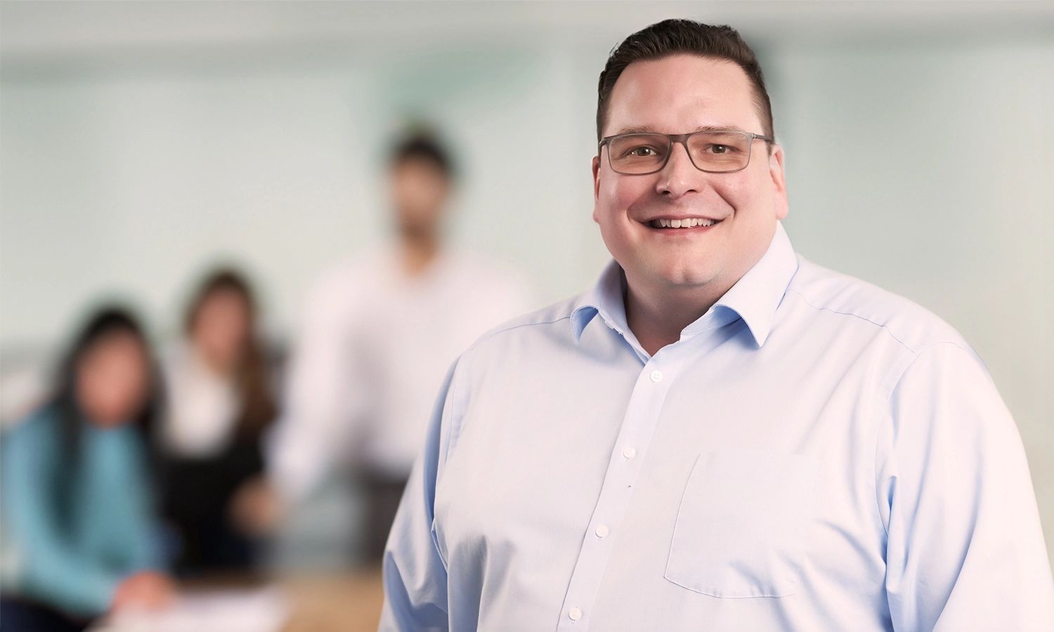 Lächelnder Schulungsleiter mit Brille steht im Vordergrund. Im Hintergrund sind weitere Teilnehmende einer Schulung zu sehen.
