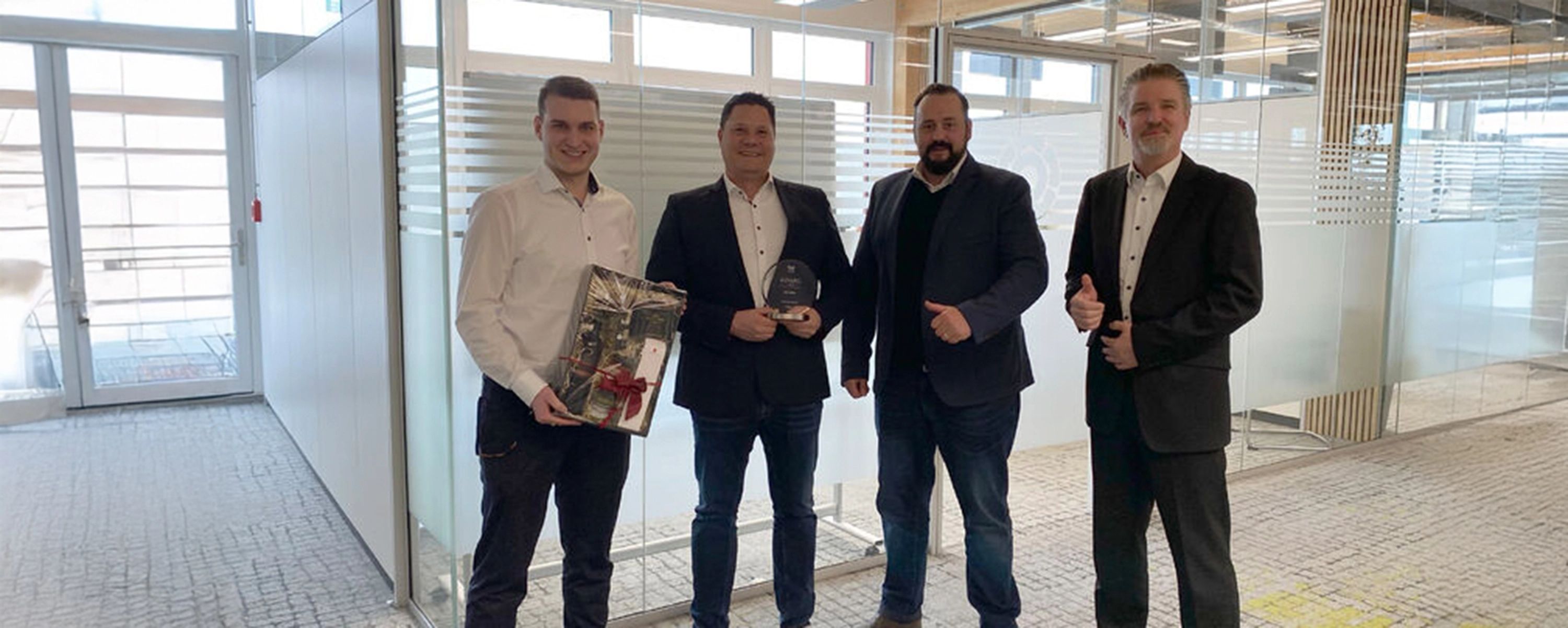 Vier Männer in Business-Kleidung posieren stolz für ein Foto in einem hellen, modernen Büro. Einer hältn anderer eine Trophäe, und zwei Männer zeigen als Geste des Erfolgs den Daumen nach oben.