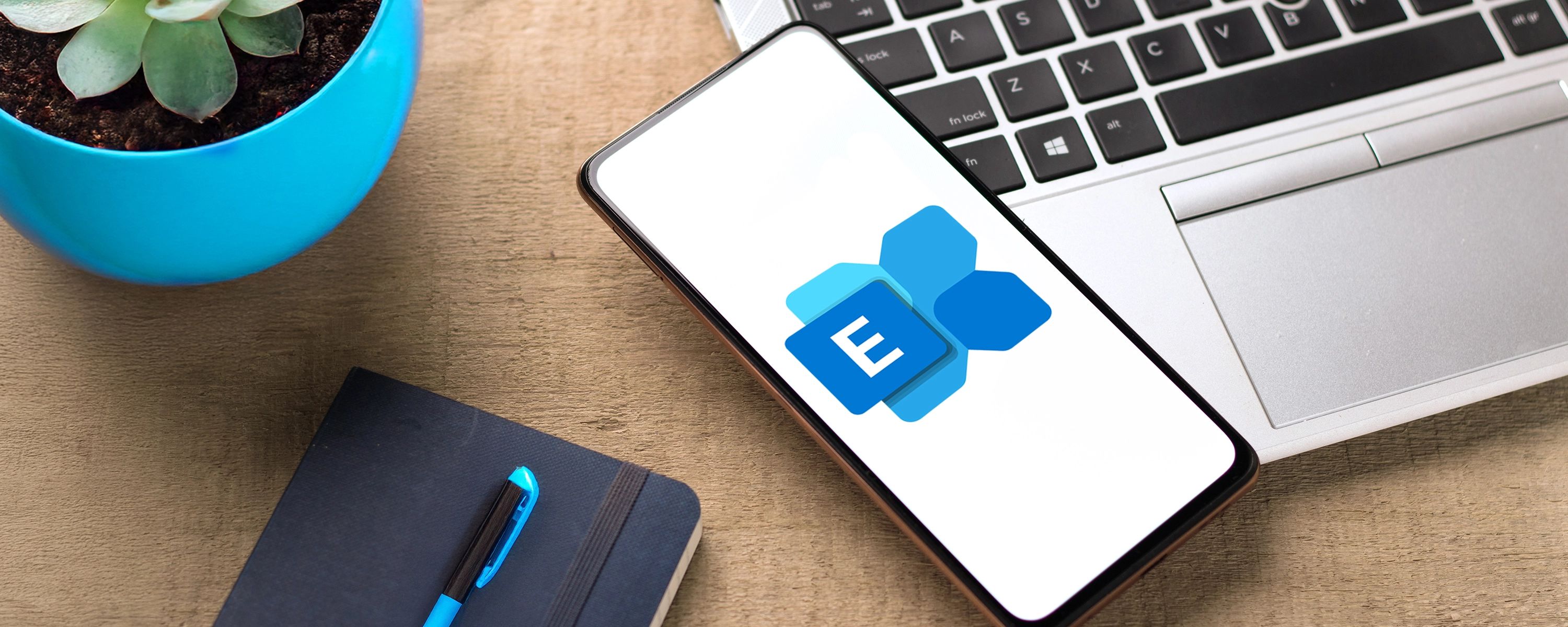 Das Logo von Microsoft Exchange wird auf einem Smartphone angezeigt, das auf einem Holzschreibtisch neben einem Laptop, einem Notizbuch und einer Pflanze liegt.