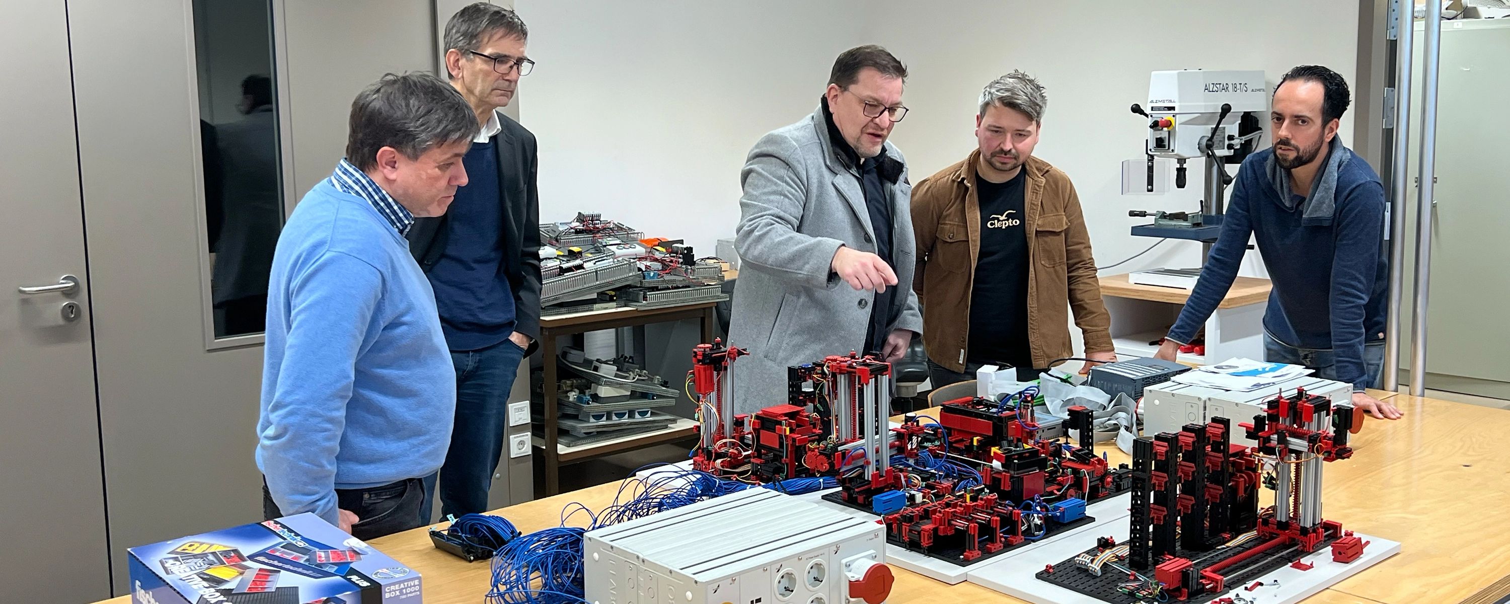 Fünf Männer in einer Werkstatt, die sich um einen Tisch versammelt haben, auf dem komplexe technische Modelle aus Bauteilen aufgebaut sind. Ein Mann in der Mitte erklärt der Gruppe gestikulierend die Funktionsweise.