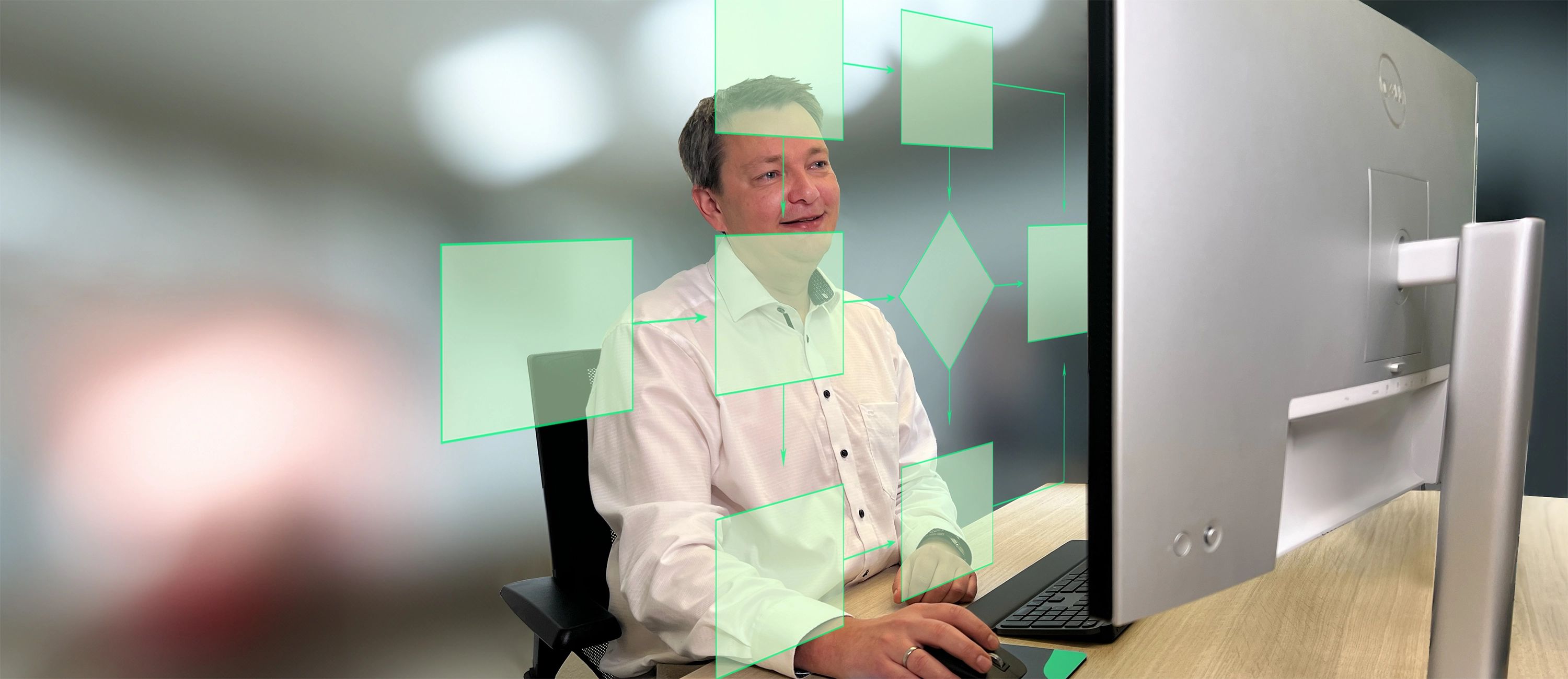 Mitarbeiter sitzt lächelnd vor dem Monitor, über dem ein leuchtendes, grünes Flussdiagramm (Workflow-Automatisierung) visualisiert wird.