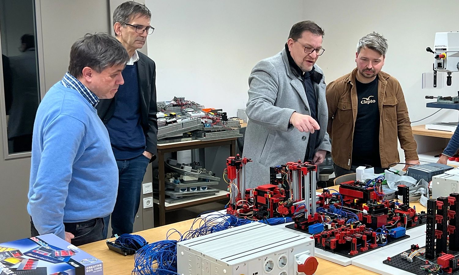 Fünf Männer in einer Werkstatt, die sich um einen Tisch versammelt haben, auf dem komplexe technische Modelle aus Bauteilen aufgebaut sind. Ein Mann in der Mitte erklärt der Gruppe gestikulierend die Funktionsweise.