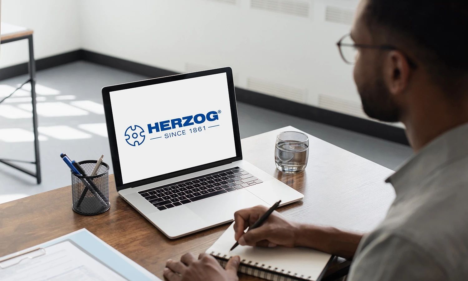 Person sitzt an einem Schreibtisch und schaut auf einen Laptop, auf dessen Bildschirm das blaue Logo von HERZOG mit dem Schriftzug ‚HERZOG – Since 1861‘ und einem stilisierten Zahnrad-Symbol angezeigt wird.