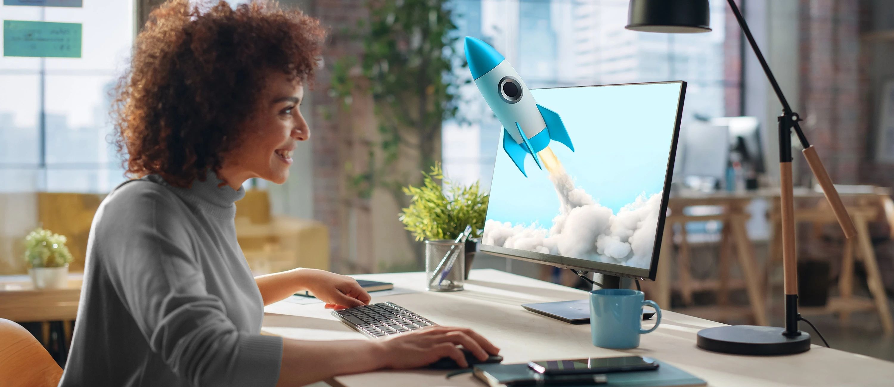 Eine lächelnde Frau mit lockigen Haaren sitzt in einem Büro vor einem Computer. Auf dem Bildschirm ist eine 3D-Animation einer blauen und weißen Rakete zu sehen, die mit viel Rauch abhebt.