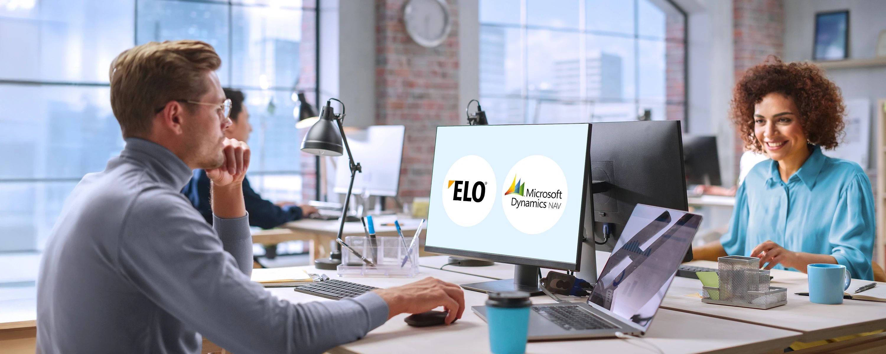 Ein Mann sitzt an einem Schreibtisch in einem modernen Büro und schaut auf einen Computermonitor. Der Bildschirm zeigt nebeneinander die Logos von "ELO" und "Microsoft Dynamics NAV". Im Hintergrund lächelt eine Frau ebenfalls im Büro.