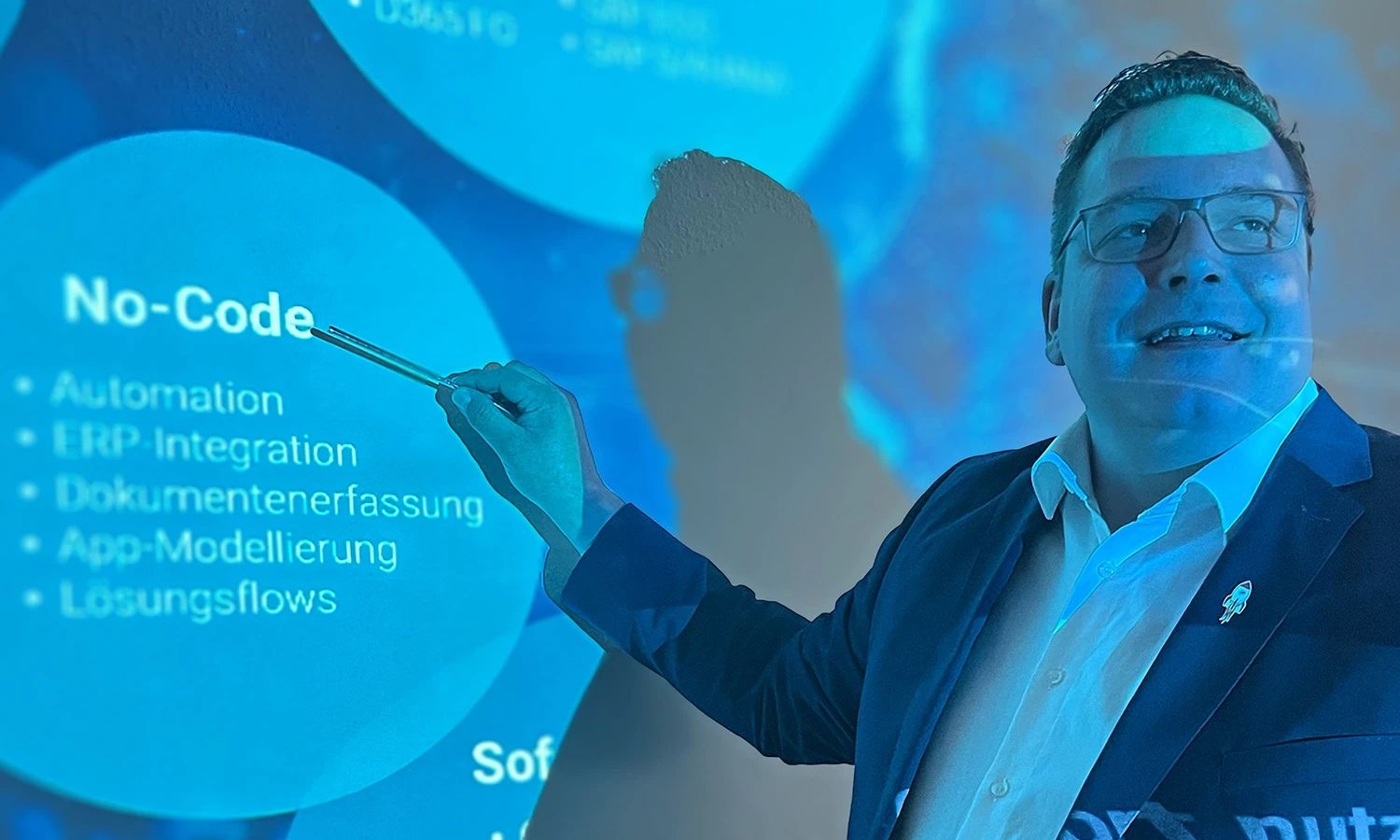 Mann hält eine Schulung ab und zeigt mit einem Stift auf eine blaue Projektion mit dem Titel "No-Code" und einer Liste von Stichpunkten.