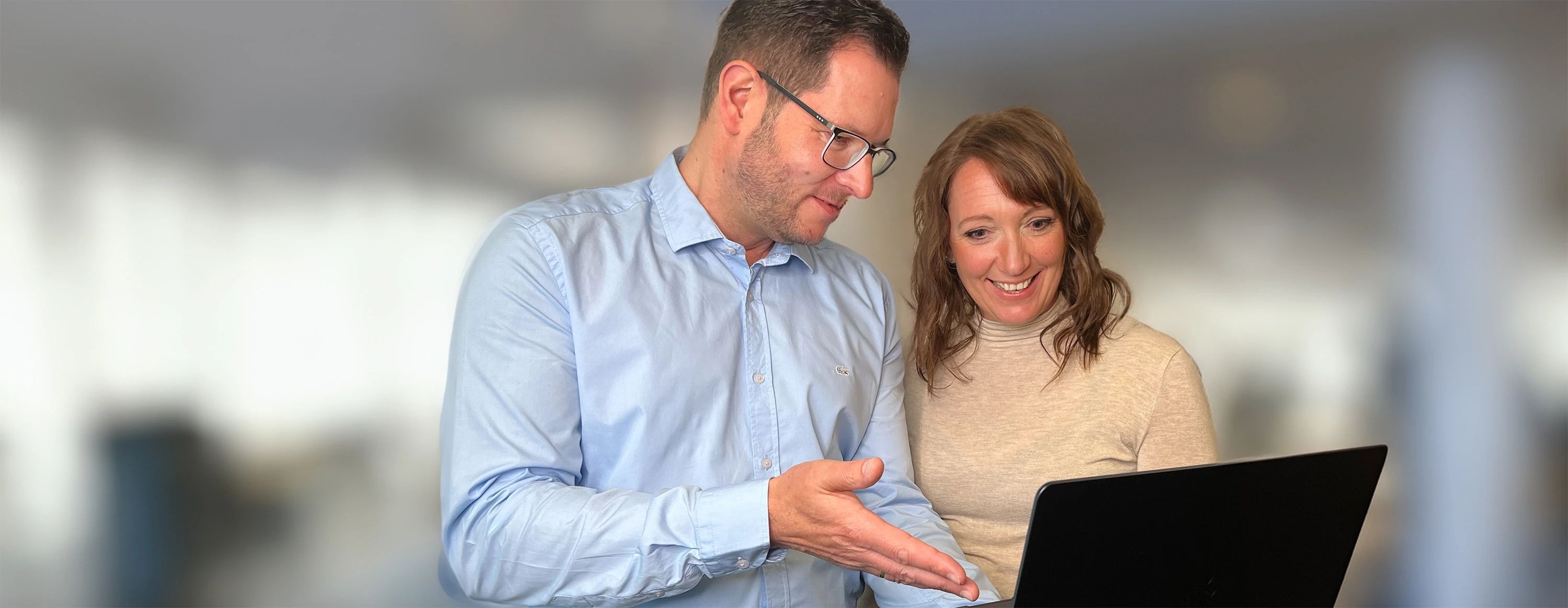 Zwei lächelnde Mitarbeitende beugen sich über einen Laptop in einer Besprechungssituation. Der Mann deutet auf den Bildschirm.
