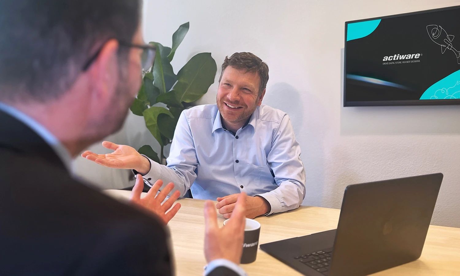 Zwei Männer in einer Besprechung, bei der einer lächelnd und gestikulierend dem anderen gegenübersitzt. Im Hintergrund ist ein Bildschirm mit dem "ACTIWARE" Logo.