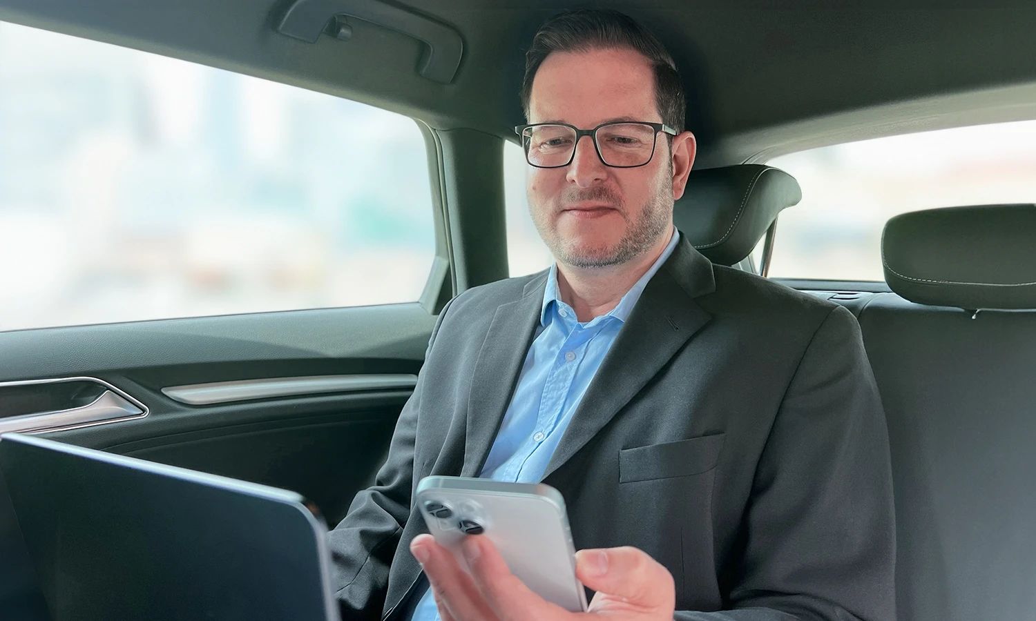 Geschäftsmann nutzt Laptop und Smartphone im Auto für mobiles Arbeiten.